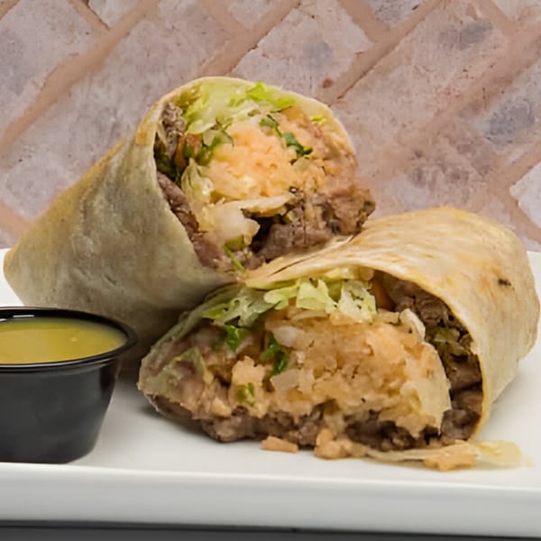 A Nuestro Sabor burrito, cut in half, revealing meat, rice, and lettuce, with a side of sauce.